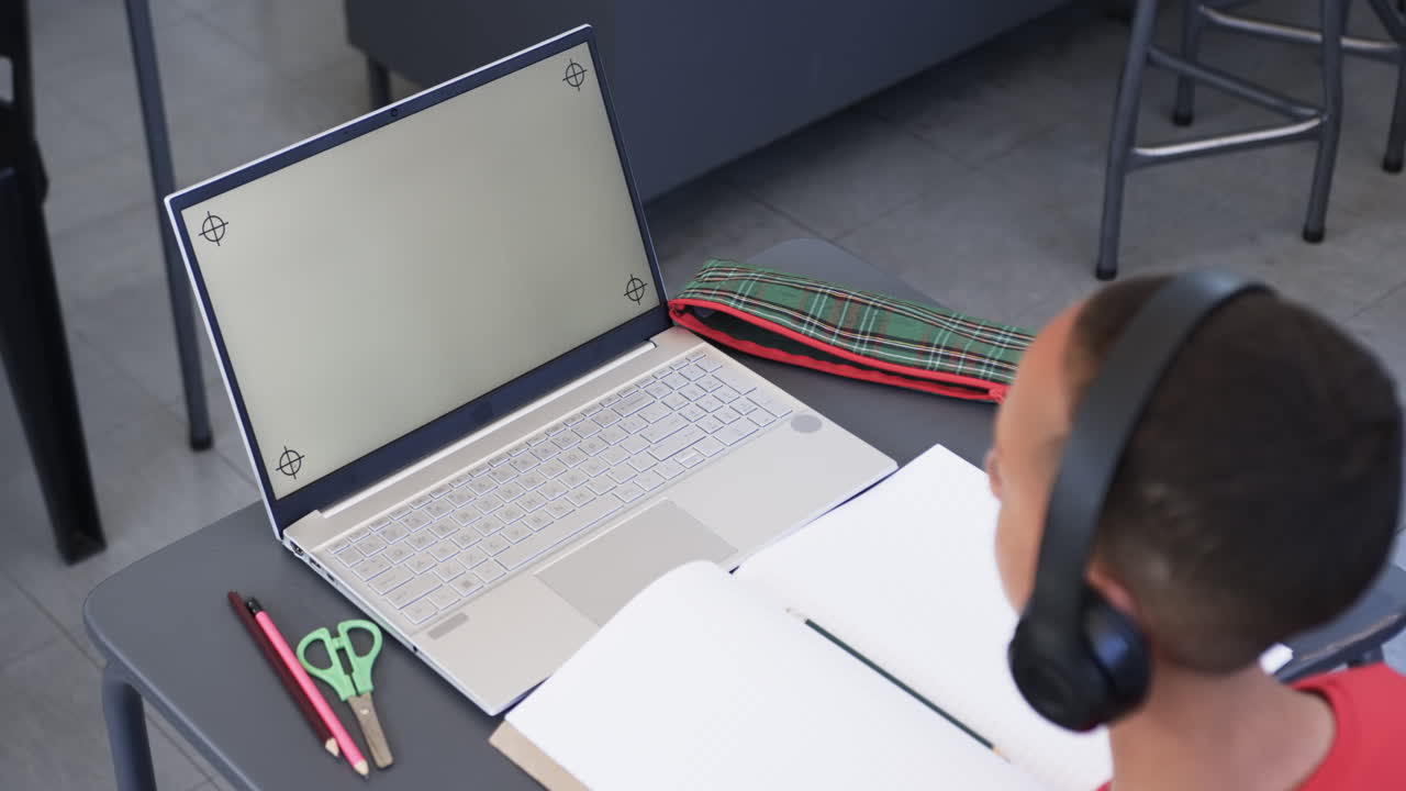 In school, biracial young boy wearing headphones focusing on laptop screen, copy space