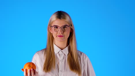 inteligente mujer joven rubia caucásica con gafas juega con un naranja feliz, fondo infinito croma azul retrato disparado