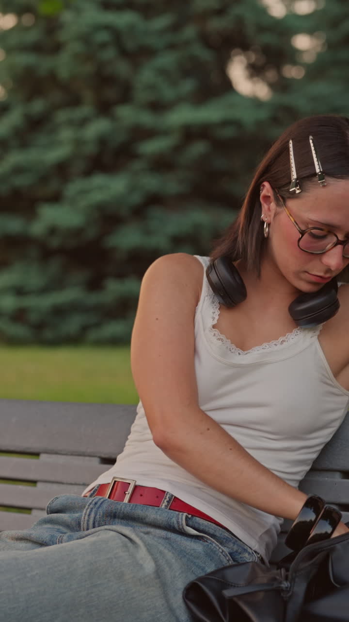 A young woman sits on a park bench, looks at nature, puts away her phone, and writes in her journal. Close-up emotional digital detox moment captured in a smooth vertical 4K shot