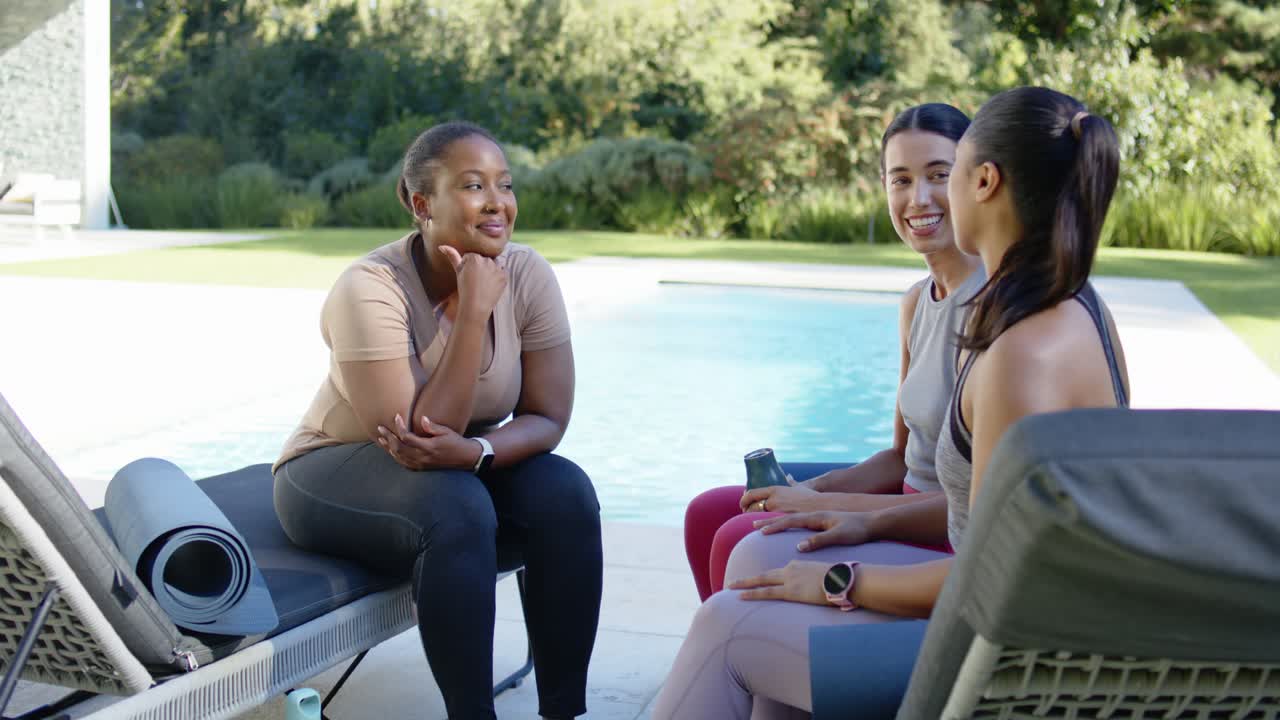 Diverse female friends chatting on poolside chairs after workout, showing yoga mat and water bottle