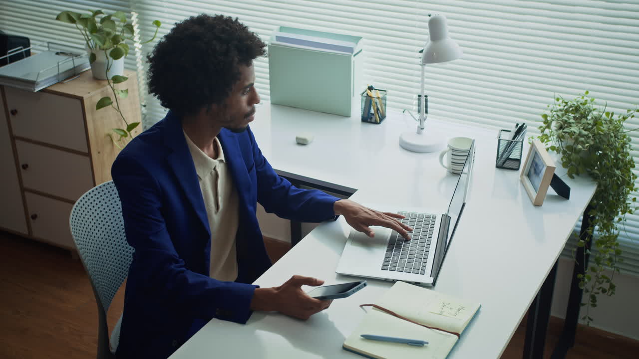 African American Businessman Checking Smartphone in front of Laptop at Office