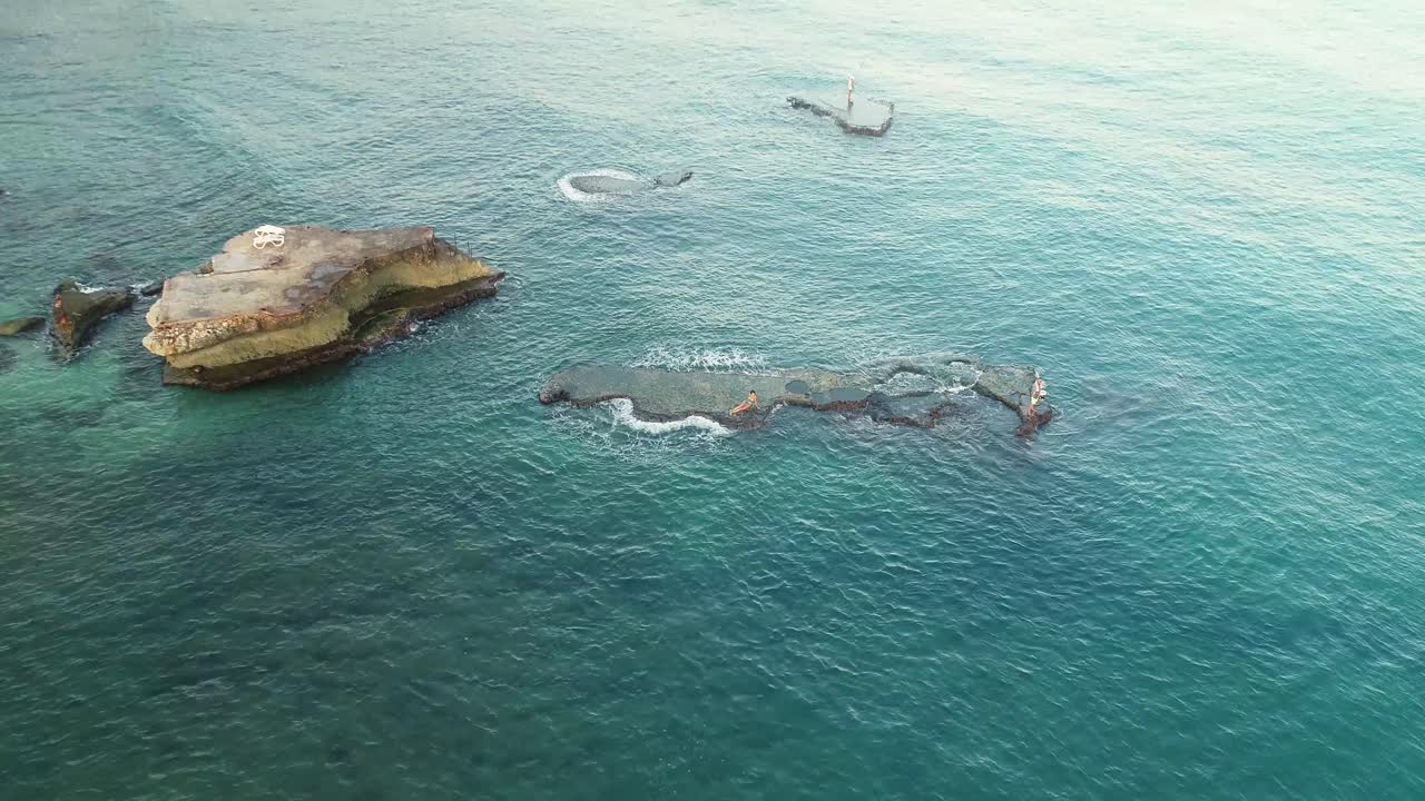 dos personas caucásicas descansando y disfrutando de la isla rocosa de batroun en un afloramiento rocoso en aguas verdes y claras del océano en un día soleado de verano, líbano, elevación aérea hacia atrás