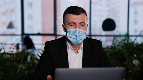 businessman wearing surgical mask works at laptop. man sitting at computer in a big office. blurred backdrop.
