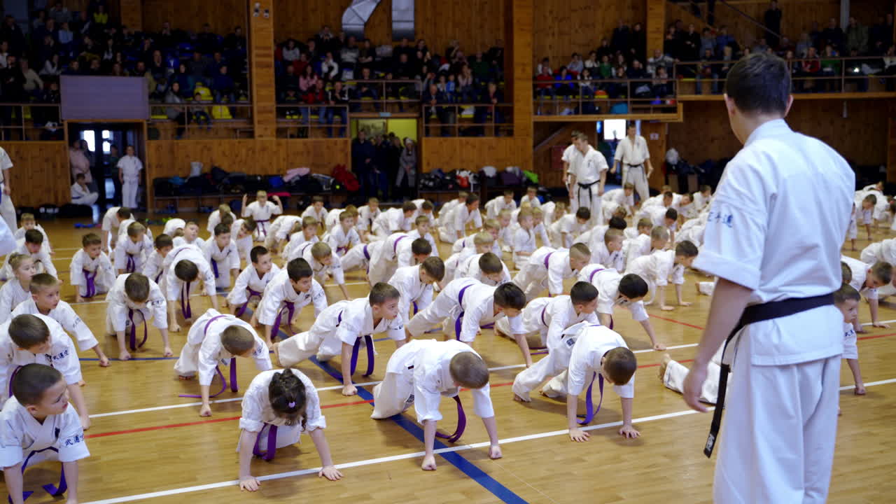 Large group of young karate athletes doing exercise on the floor. Numerous spectators watch the trainees sitting at backdrop.