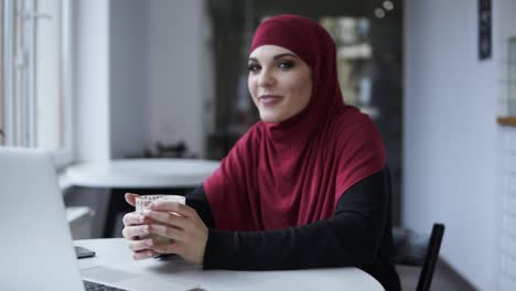 Attractive-young-shy-female-in-hijab-drinking-cappuccino-in-some-minimalistic-cafe.-Indoors-footage