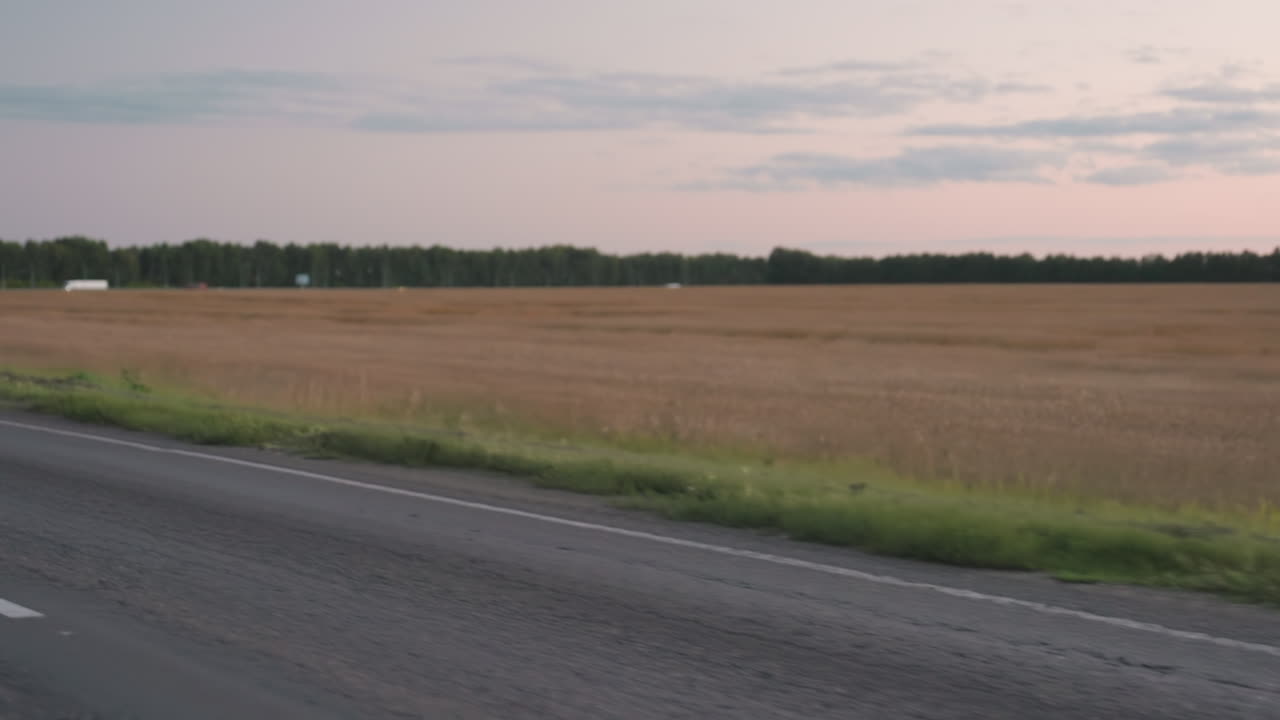 Motorbike rider wearing helmet travels along countryside road with passenger, headlight glowing as asphalt stretches past golden farmland fields and tree line horizon, evening sky