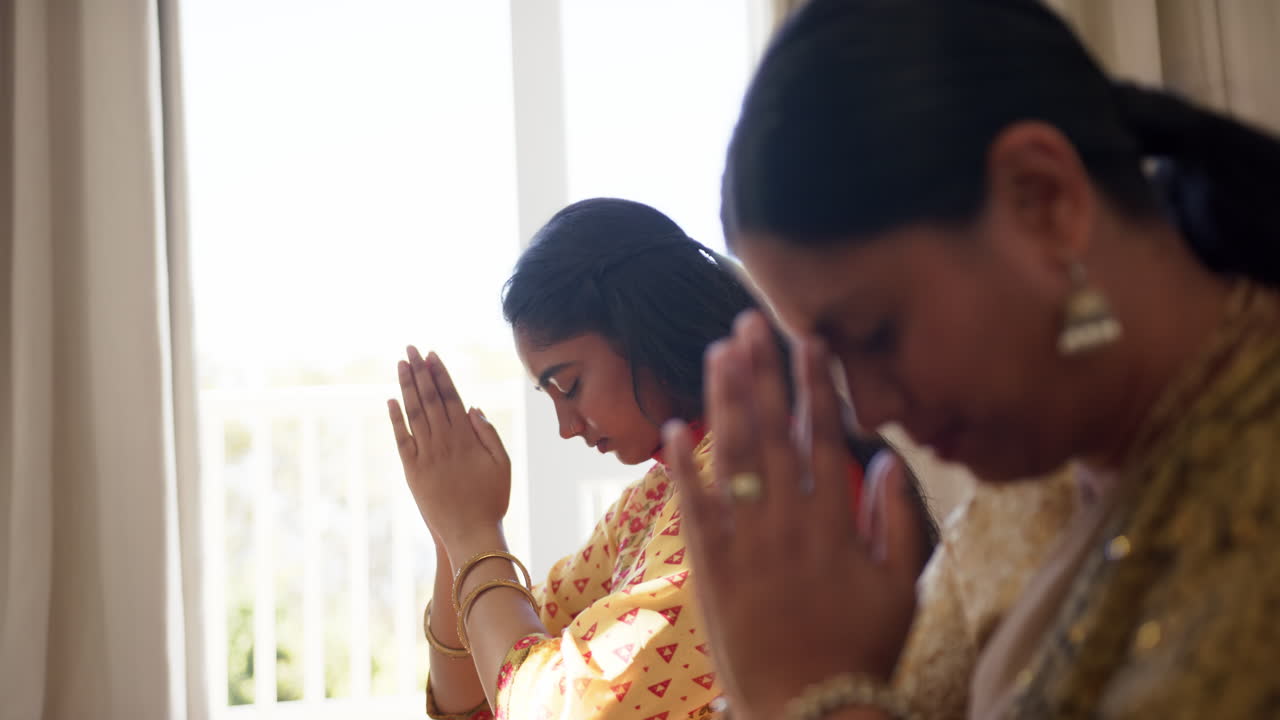 Praying together, indian mother and daughter bowing heads and holding hands in traditional sari