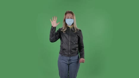 의료 보호 얼굴 마스크를 입은 아름다운 젊은 여성의 초상화는 카메라를보고 주먹을 보여 주고, 한, 두, 세, 네, 다섯 손가락을 세고, 검은색 재과 청바지를 입은 느슨한 머리카락을 가진 금발은 스튜디오에서 녹색 화면에 건강 보호.
