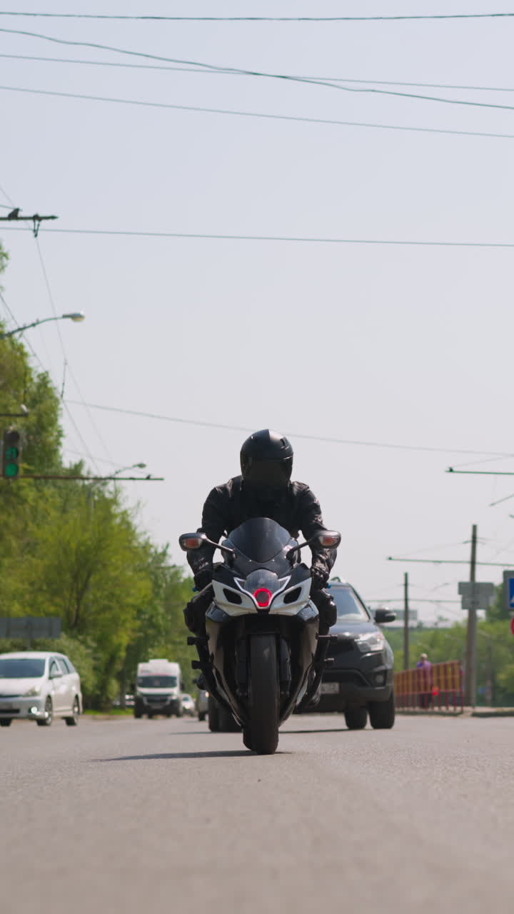 Man driver in black leather jacket and helmet rides powerful bike along large road with cars in modern city on sunny day front view slow motion