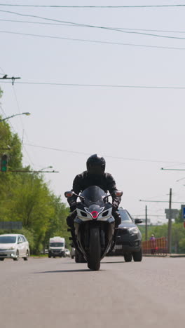 man driver in black leather jacket and helmet rides powerful bike along large road with cars in modern city on sunny day front view slow motion