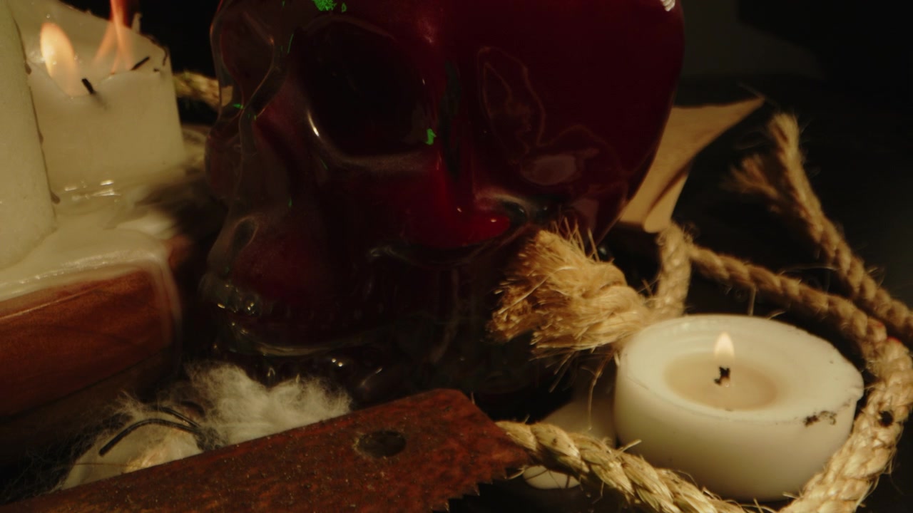 Concetto horror occulto: macro ripresa in allontanamento di un teschio di vetro pieno di sangue sull'altare di una strega con corde insanguinate, candele accese e vetri rotti.