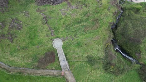 Aerial-birdseye-shot-over-the-Gobbins-Cliffs-in-beautiful-Northern-Ireland-overlooking-the-rocky-slope-in-front-of-the-blue-sea-with-rocks-in-the-water-during-an-adventurous-journey