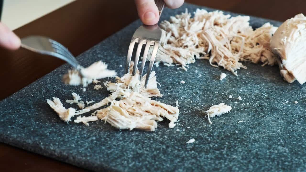 A man slicing boiled chicken breast on a cooking board using two forks