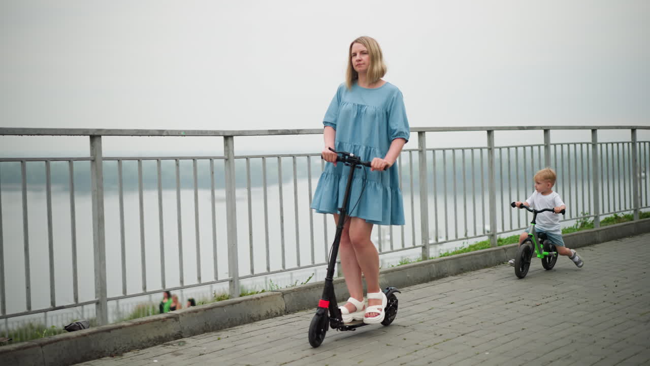 A woman rides a scooter with a thoughtful expression, followed by a young boy on a bicycle, they move along a bridge, with a scenic water body in the blurred background