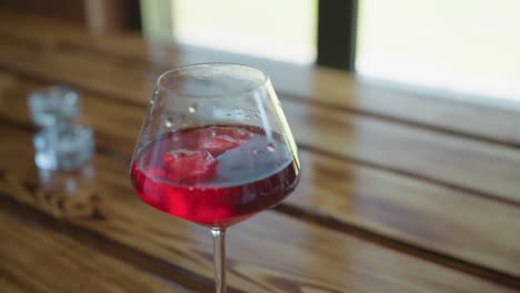 closeup hand gently drops ice cube into elegant wine glass filled with red drink resting on polished wooden table as natural light filters softly through window creating cozy serene ambiance indoors