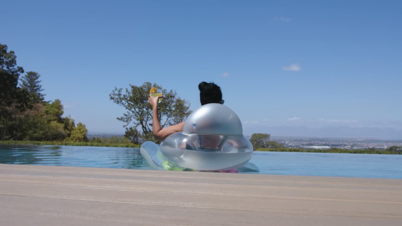Mature woman holding yellow cup sitting in float at infinity pool in bikini, trailing hand, basking