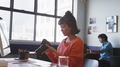 Biracial-businesswoman-sitting-at-table-and-inspecting-camera-at-office