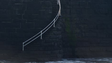 waves crash over steps on a dark coloured stone brick sea wall with white railings