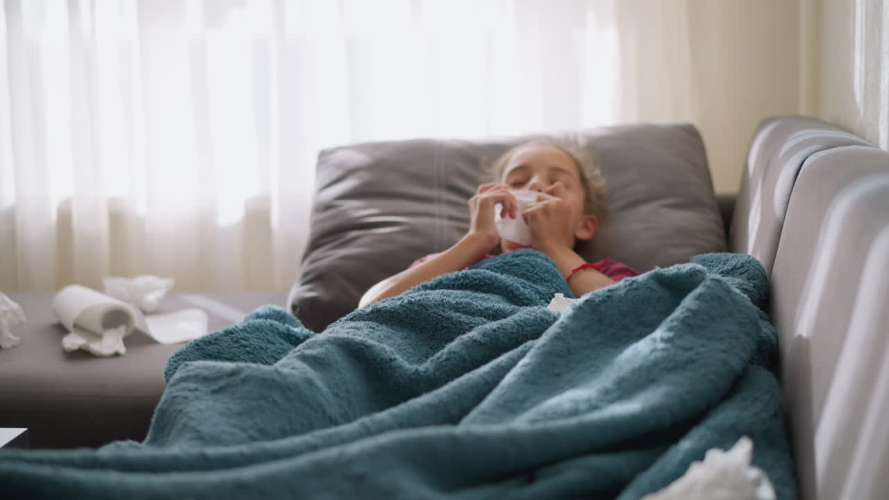 Ragazza bianca sdraiata sul divano avvolta in una coperta, con fazzoletti usati sparsi intorno a lei, a indicare che è malata e si sta riprendendo da un raffreddore o un'influenza, con una zona circostante disordinata di fazzoletti.