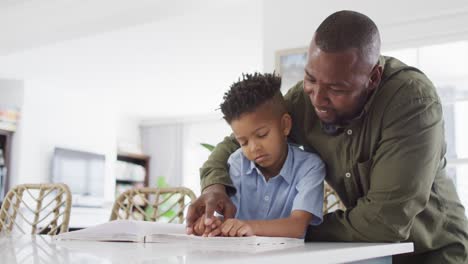 Happy-african-american-man-and-his-son-sitting-at-table-and-reading-braille