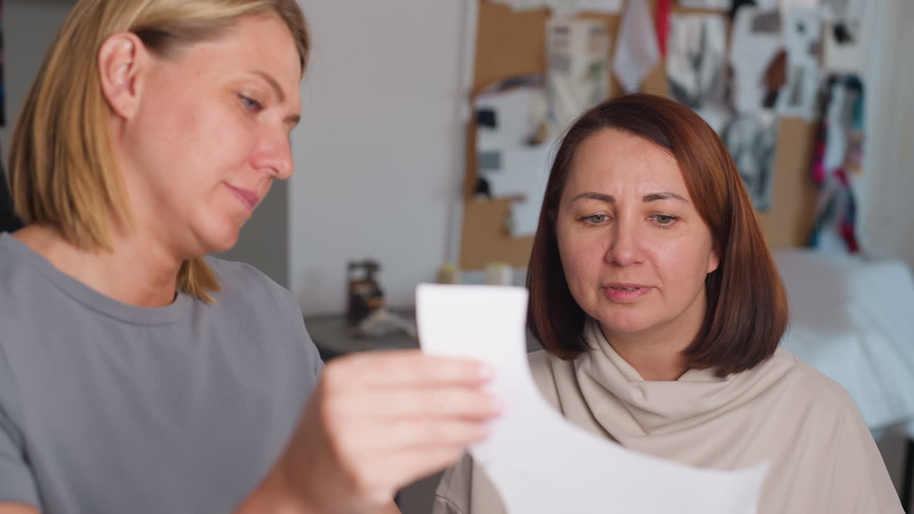 Costumer holding paper pattern showing it to friend making funny face while smiling warmly, both engaging in lighthearted discussion in workshop environment surrounded by design inspiration