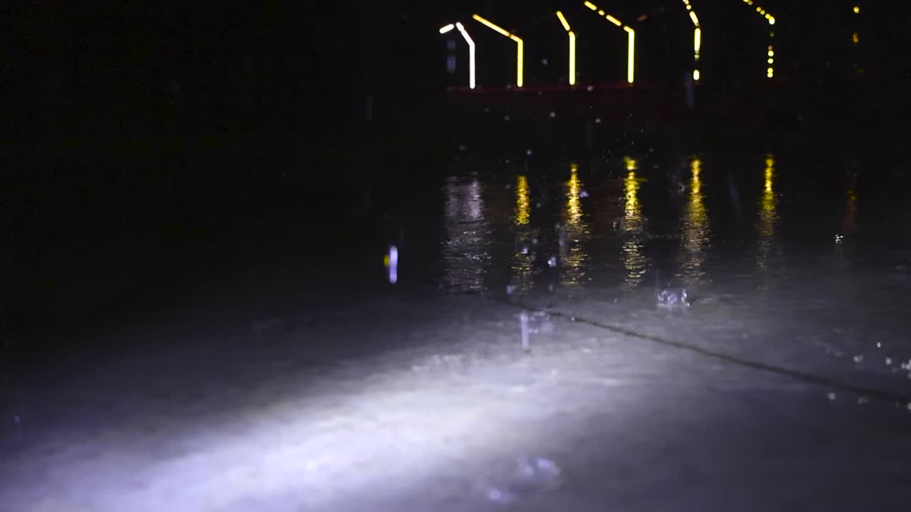 Low angle video of heavy and large raindrops falling on a gray concrete floot outdoors in the night time in slow motion, with water splatter and splashing. Design LED lights at bokeh dark background