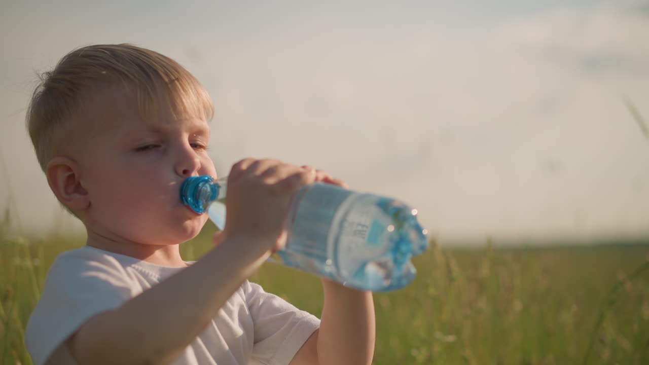 A little kid wearing a white top is sitting in a grassy field, putting a water bottle to his mouth to drink. The child appears focused and calm, enjoying a refreshing moment outdoors in the sunlight