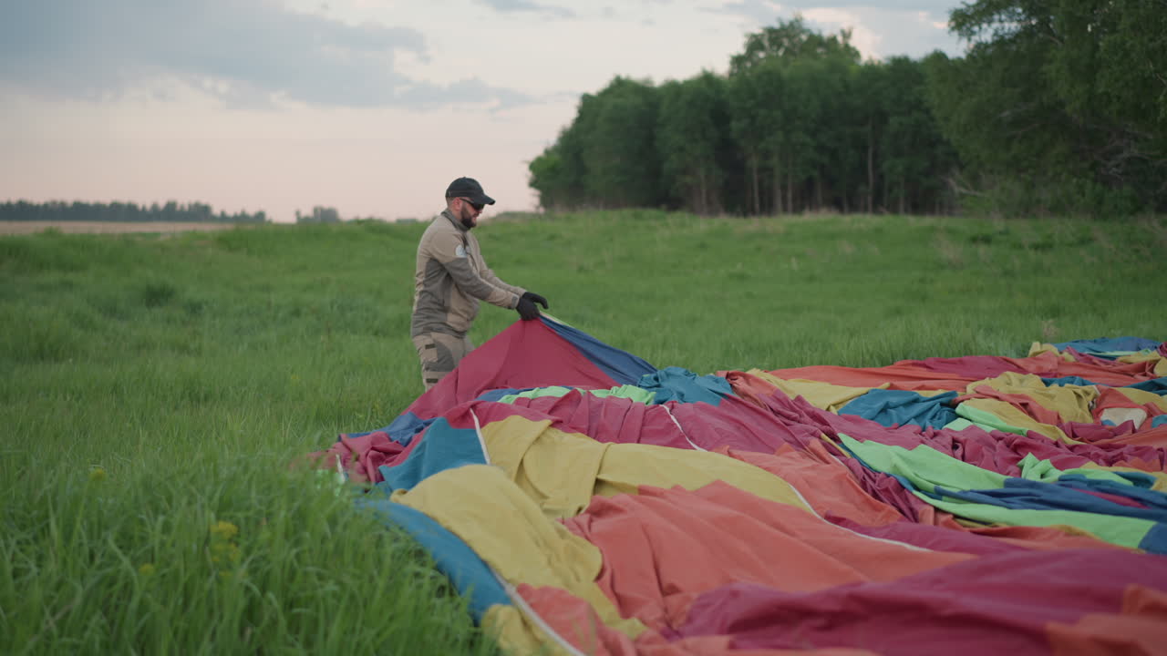 man in dark cap and gloves pulling colorful balloon fabric across grassy meadow near forest edge under pastel sky arranging material for hot air balloon launch during golden hour
