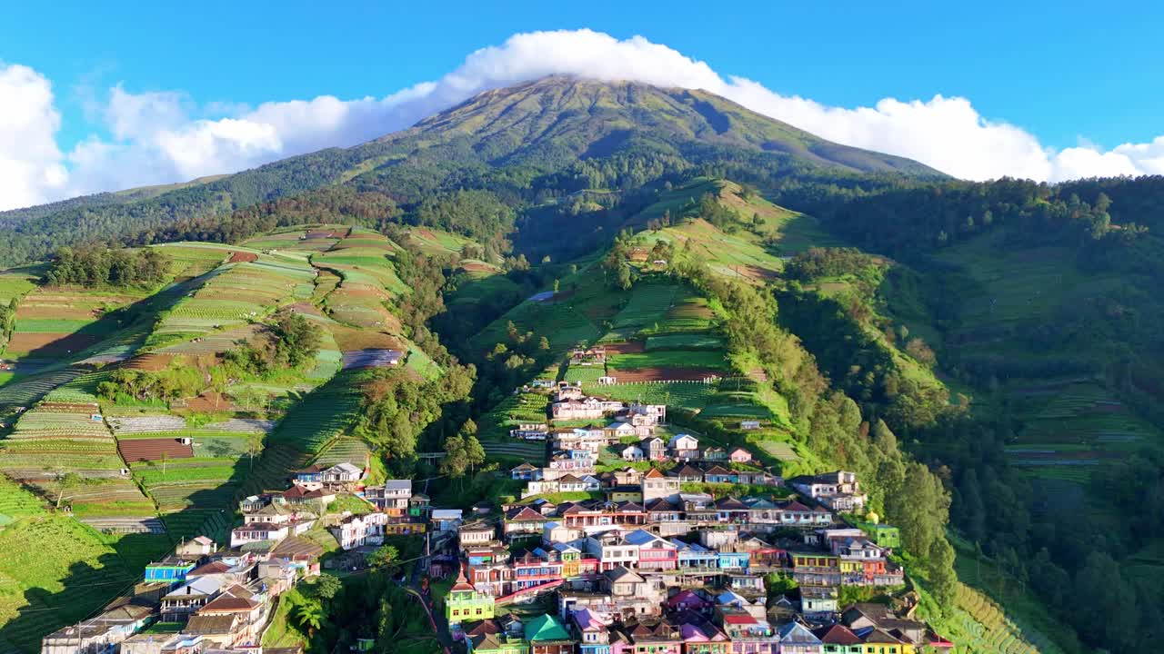 Drone footage of Nepal van Java, a vibrant mountain village in Central Java, Indonesia. The scene showcases colorful houses stacked along steep hillsides beneath Mount Sumbing’s majestic peak