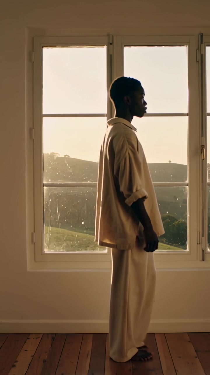 Vertical video: Placing right foot, man walking across room loose shirt, window backlit to cross