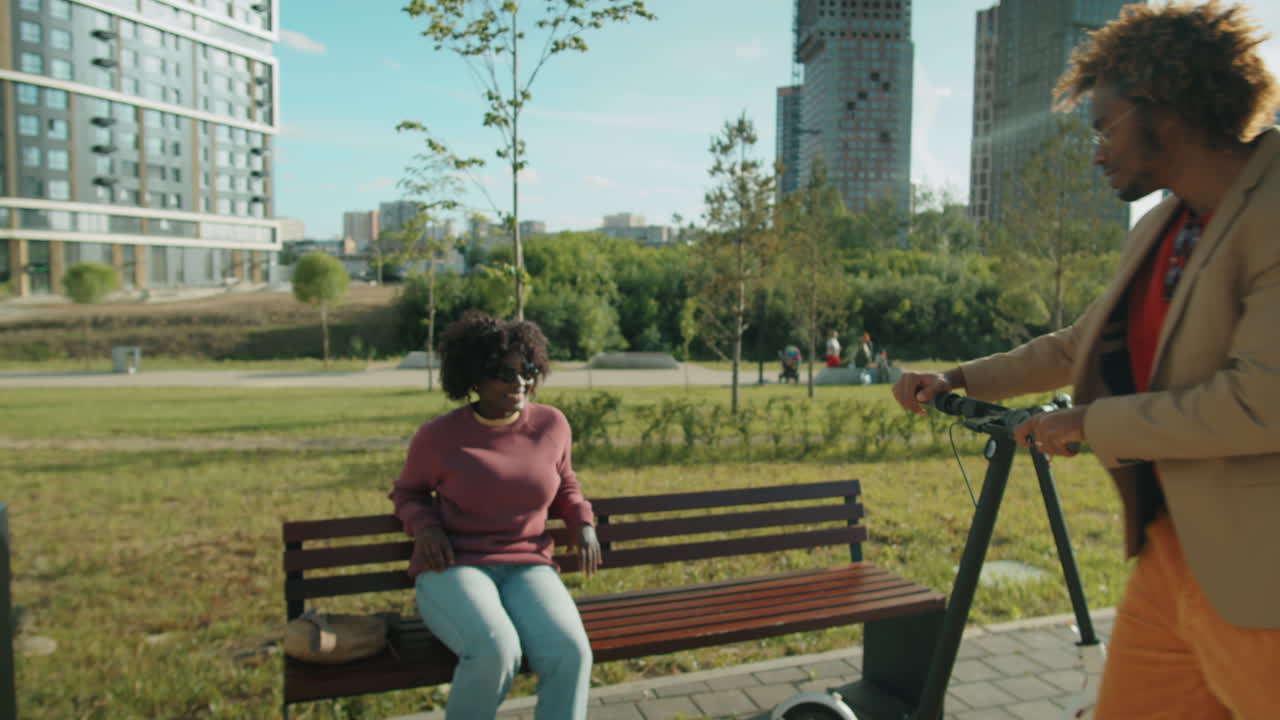 hombre afroamericano montando un scooter eléctrico en el parque y charlando con su novia en el banco