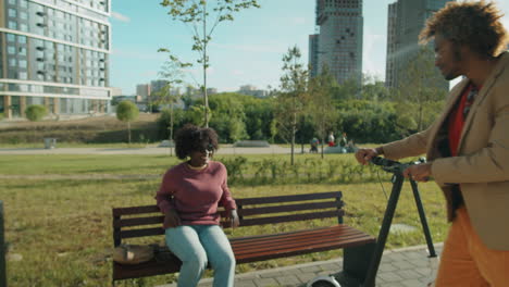 african american man riding e-scooter in park and chatting with girlfriend on bench