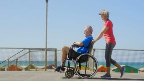 side view of active senior caucasian woman pushing senior man on wheelchair at beach 4k