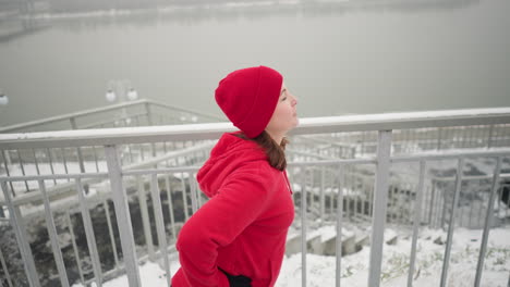 mujer de negocios haciendo ejercicio al aire libre durante el invierno, sosteniendo una barandilla de hierro para el equilibrio con una cálida sonrisa, rodeada de tierra nevada, atmósfera brumosa, río, puente y postes de luz en el fondo