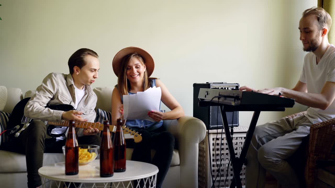 una atractiva vocalista y un alegre guitarrista están leyendo letras de canciones y riendo mientras el tecladista está tocando el teclado y ajustando el instrumento musical.