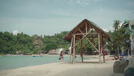 Los-Turistas-Disfrutan-Caminando-Por-La-Playa-En-El-Nido,-Filipinas-Con-árboles-Exóticos-Y-Cabañas-De-Madera---Perfecto-Para-Las-Vacaciones-De-Verano---Plano-General