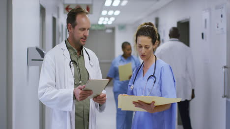 Médico-Masculino-Y-Femenino-Caucásico-Hablando-En-El-Pasillo-Del-Hospital-Mirando-Tabletas-Y-Archivos-De-Pacientes