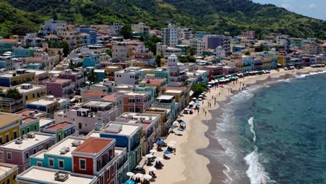 aerial video captures vibrant coastal town with colorful buildings, sandy beach, and lush hills