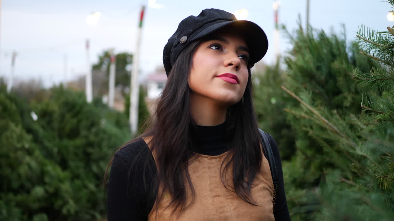 una mujer hispana comprando un árbol de navidad de temporada en un lote con muchas especies de árboles festivos