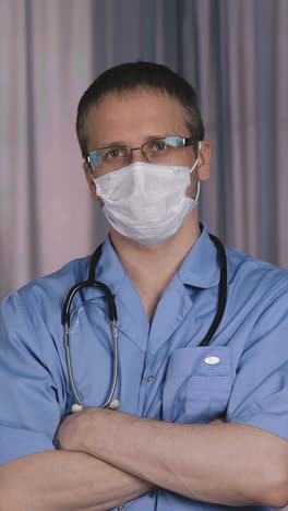 portrait of young doctor, with stethoscope around his neck, wearing protective mask, glasses, looking into camera, on background in his office, fold one's arms