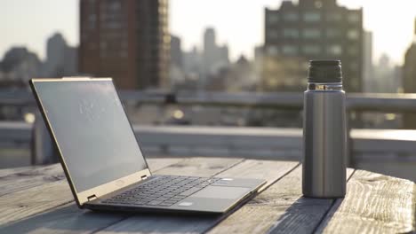 a tranquil workspace scene showing a laptop and a stainless steel thermos on a wooden table with a cityscape backdrop during golden hour, emphasizing productivity and comfort