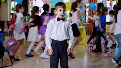 lonely kid wearing bow tie standing motionless, watching blurred children playing during festive birthday celebration, expressing isolation and melancholy
