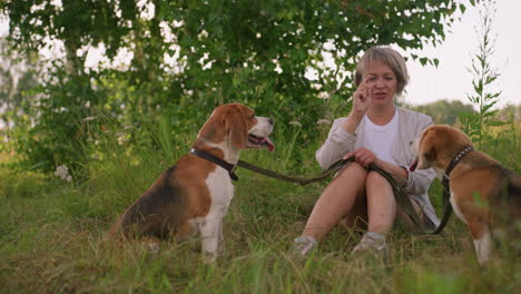 pet lover sitting outdoors in grassy field interacting with her dogs on leash, playfully teasing one with hand gesture as dog on left moves closer while second dog watches with tongue out