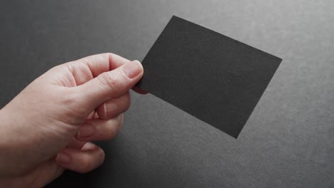 Hand-of-caucasian-woman-holding-black-business-card-on-grey-background,-copy-space,-slow-motion