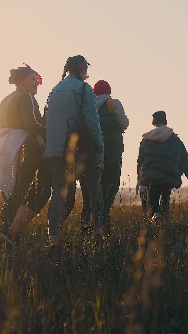 group of young people walk along meadow to cars after day hike in autumn evening backside view