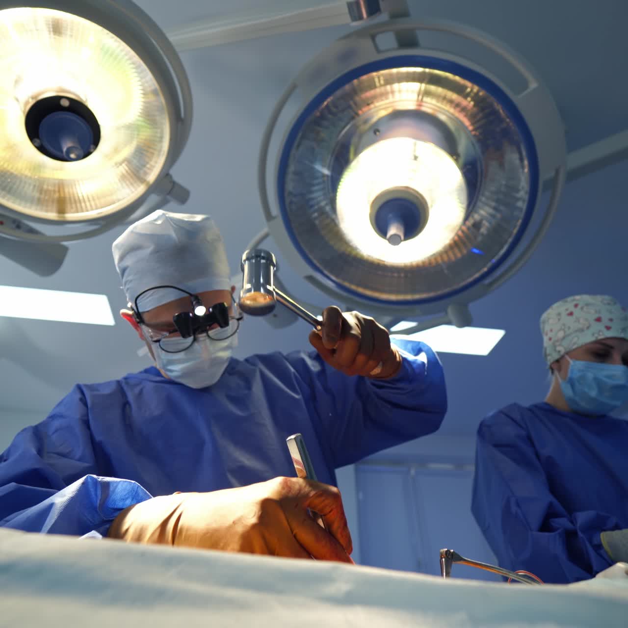 Surgeon uses special surgical tools during operation. Professional doctor and a nurse in medical uniform on bright lamps background in clinic. View from below