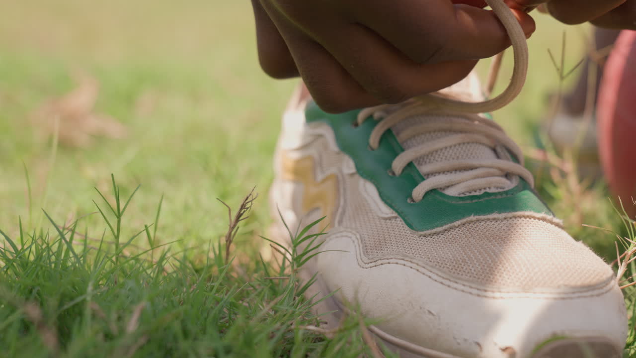 Bambino si allaccia le scarpe su un campo erboso. Giovane bambino che lega le scarpe da ginnastica vicino a un vivace pallone rosso all'aperto. Ragazzo che allaccia con cura i suoi lacci delle scarpe accanto a un pallone rosso brillante nel parco sotto la calda luce del sole.