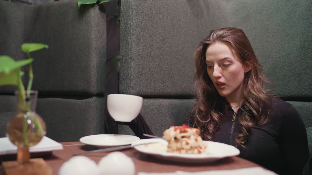 Lady in black top gently sips latte while seated in cozy booth and slowly returns cup to white saucer, with creamy waffles, green plant in vase, and soft indoor lighting creating warm ambiance