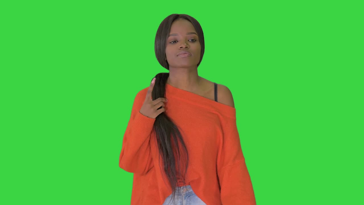 Donna afroamericana sorridente vestita con un maglione luminoso e jeans, che cammina toccandosi i capelli su uno schermo verde, Chroma Key