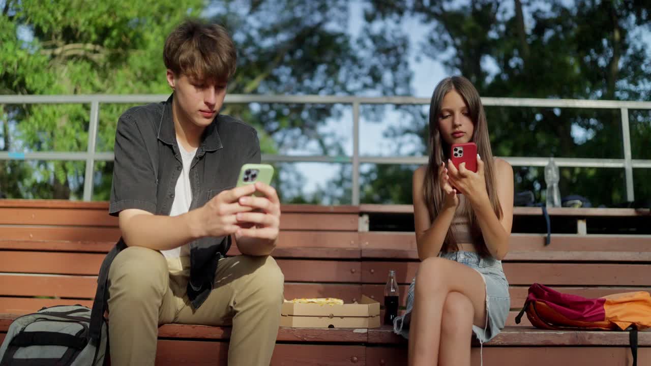 una pareja joven sentada en un banco mirando sus teléfonos.